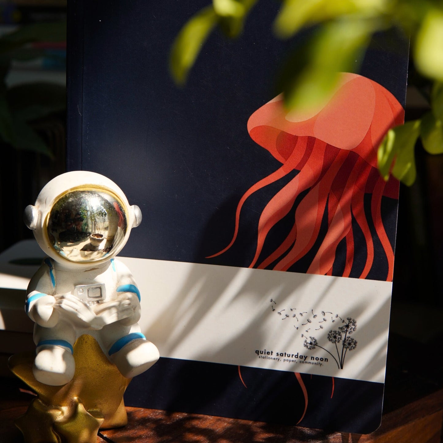 Small astronaut figurine next to a journal of a jellyfish on a wooden surface with plants in the background.